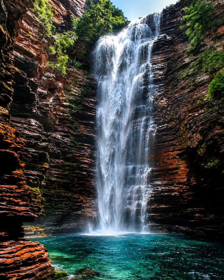 Cachoeira caindo por um paredão de rocha ocre coberto de mata, formando ao pé um poço de água turquesa