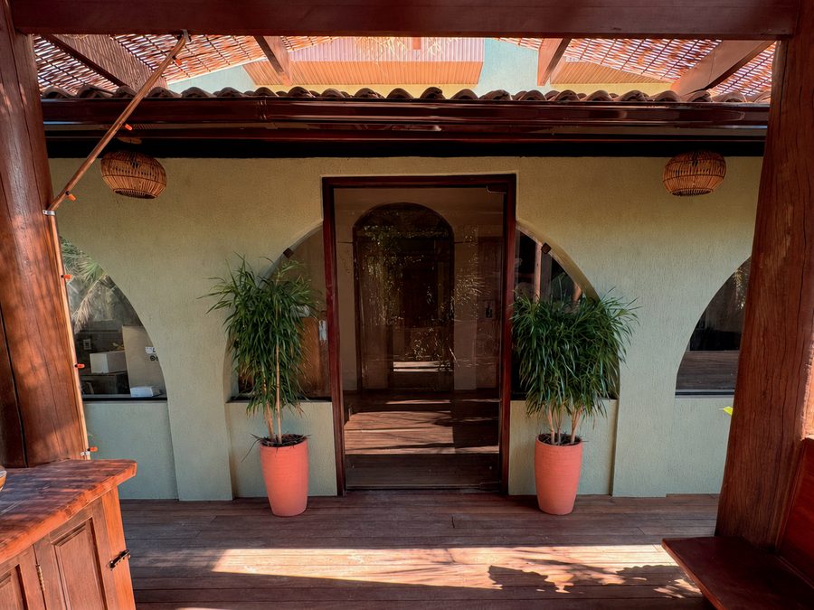 Varanda de entrada do Instituto: vigas e deque de madeira sob pergolado de bambu, luminárias de fibra, vasos com plantas e portal de vidro entre arcos